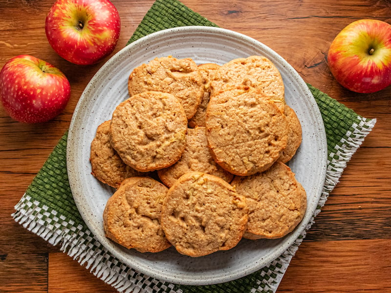 Plate of chewy apple cider cookies on a rustic wooden table, surrounded by fresh apples and a green woven napkin.