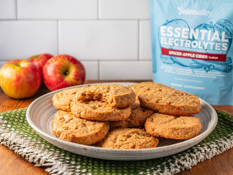 Plate of chewy apple cider cookies with a bite taken out, surrounded by fresh apples and PaleoValley Essential Electrolytes Spiced Apple Cider flavor.