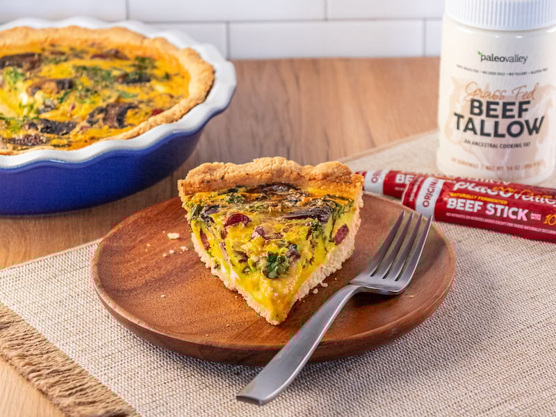 A slice of tallow-crusted quiche with mushrooms, spinach, and Paleovalley beef sticks served on a wooden plate with a fork.