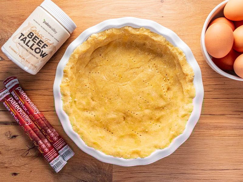 Unbaked tallow pie crust in a white dish, surrounded by Paleovalley beef sticks, beef tallow jar, and a bowl of fresh eggs.
