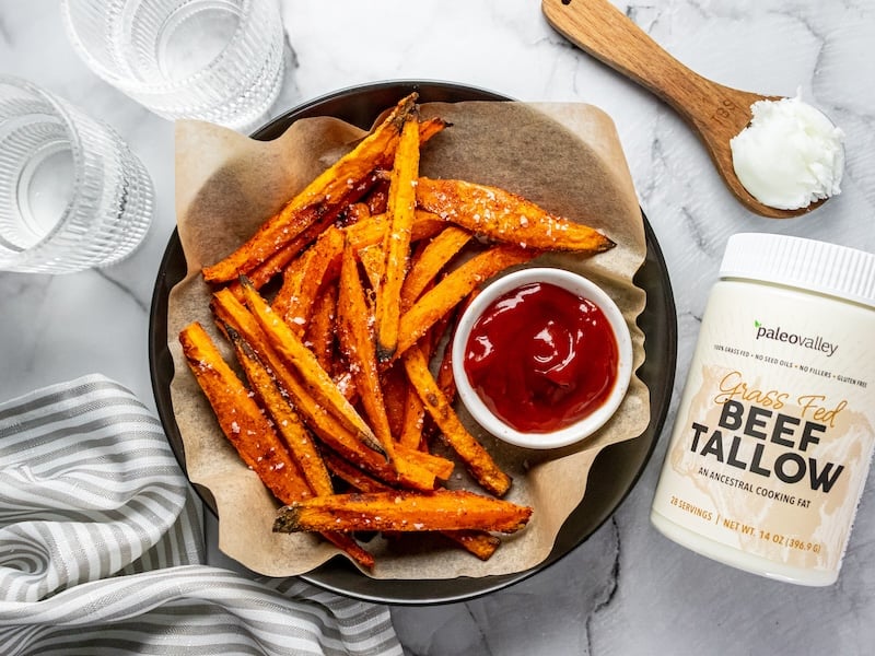 Crispy sweet potato fries cooked in Paleovalley Grass-Fed Beef Tallow, served with ketchup alongside a jar of tallow, a wooden spoon with tallow, striped towel, and glasses on a marble surface.