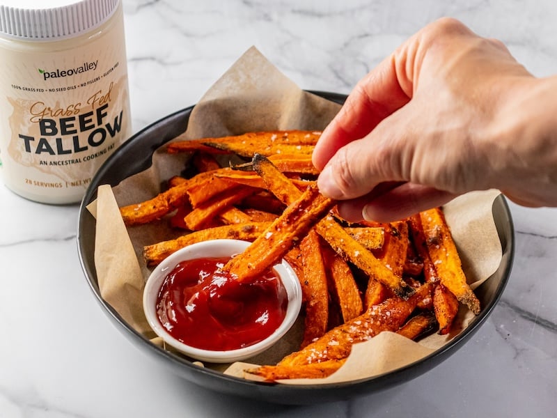 Hand dipping a crispy sweet potato fry cooked in Paleovalley Grass-Fed Beef Tallow into ketchup, with a jar of tallow in the background.